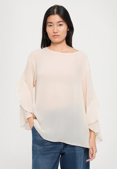 Helle beigefarbene Bluse aus durchscheinendem Stoff mit weiten, gerüschten Ärmeln und einem runden Halsausschnitt, kombiniert mit dunkellblauen Jeans.