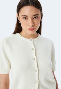 Cardigan corto crema con maniche corte e scollatura rotonda, caratterizzato da una chiusura a bottoni e delicati dettagli a coste sulle maniche e sul colletto.