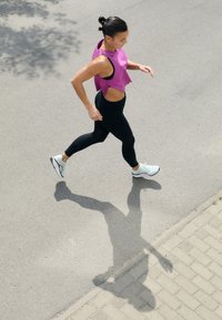 Kobieta w fioletowym topie bez rękawów i czarnych legginsach biega po utwardzonej powierzchni, nosząc wielokolorowe buty do biegania. Widoczny cień.