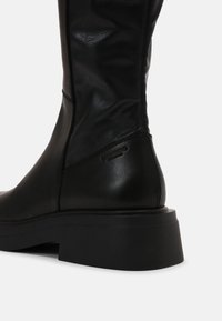 Bottine noire en cuir, hauteur genou, avec une finition texturée, dotée d'une semelle épaisse noire et d'une discrète marque sur le talon.