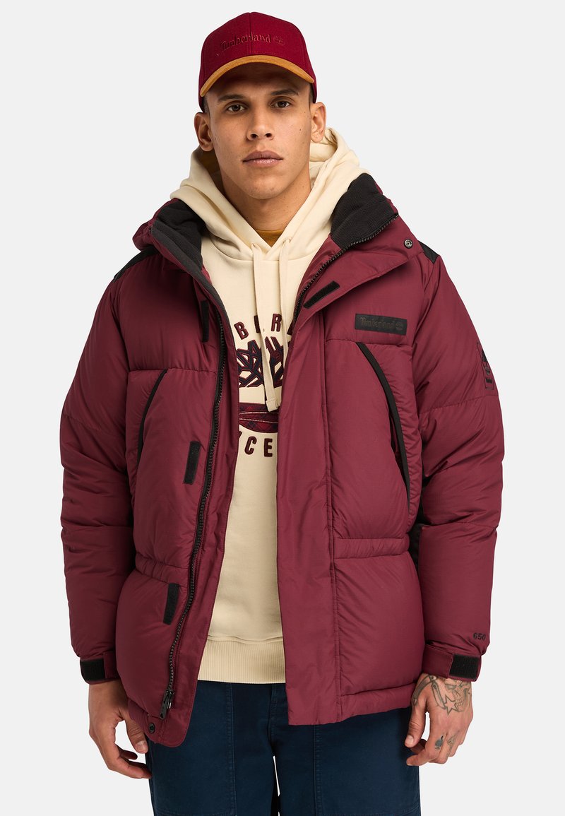 Chaqueta acolchada burdeos con detalles en negro, que presenta un cierre con cremallera en la parte delantera, bolsillos en los laterales y un cuello alto. Llevada sobre una sudadera beige.