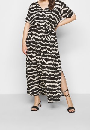 Kobieta w maxi sukience w czarno-biały wzór zygzaków, z krótkimi rękawami, rozcięciem po boku oraz czarnych sandałach na obcasie z paseczkami.