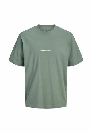 Chłopiec stojący, ubranego w czerwoną koszulę w kratę na zielony t-shirt, luźne czarne dżinsy i beżowe buty na jednolitym białym tle.