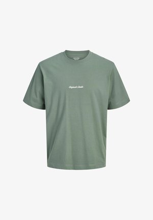 Chłopiec stojący, ubranego w czerwoną koszulę w kratę na zielony t-shirt, luźne czarne dżinsy i beżowe buty na jednolitym białym tle.