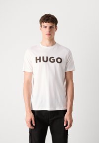 Fehér pamut póló, amelynek elején merész, sötét "HUGO" felirat található. Rövid ujjú és kerek nyakú. Minimális design.