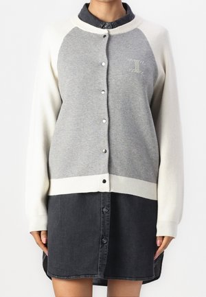 Femme portant un cardigan gris et blanc boutonné sur une robe chemise noire boutonnée, debout devant un fond blanc uni.