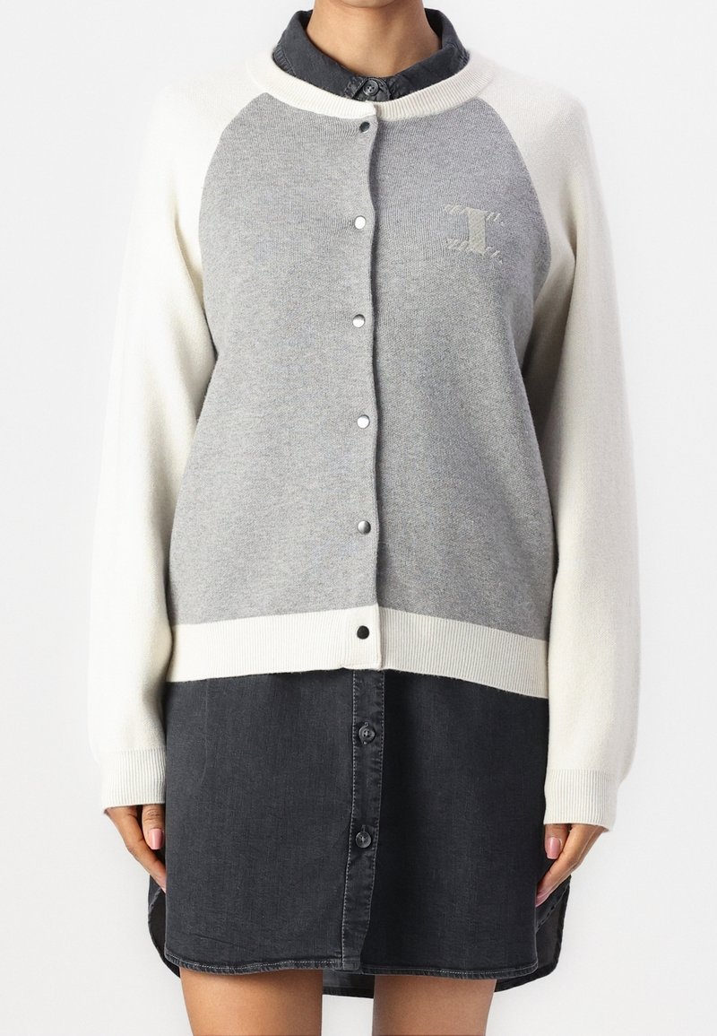 Femme portant un cardigan gris et blanc boutonné sur une robe chemise noire boutonnée, debout devant un fond blanc uni.