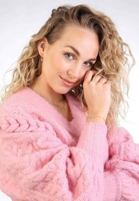 Roze kabelgebreide trui met lange mouwen, uiterlijk zacht; het onderwerp draagt delicate sieraden en heeft los, krullend haar.
