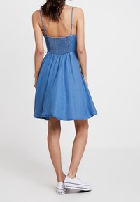 Denimklänning med spaghettistremmar, smockat midja och utsvängd kjol. Har fransade avslut. Bärs med vita sneakers.