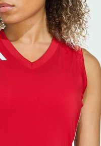 Haut de sport rouge sans manches avec un col en V, en tissu lisse et léger. Présente un logo blanc contrastant sur l'épaule gauche.