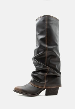 Høy svart skinnslouchy-støvel med brune detaljer, spiss tupp og stablet hæl mot en ensfarget hvit bakgrunn.