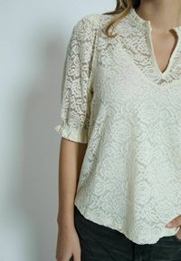 Krajková blůza v off-white barvě s výstřihem do V a krátkými nařasenými rukávy. Obsahuje složité květinové vzory a volný, texturovaný design.