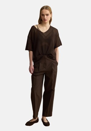 Mujer de pie con top suelto marrón oscuro semitransparente con escote en V, pantalones marrones de talle alto y bailarinas oscuras contra un fondo liso.