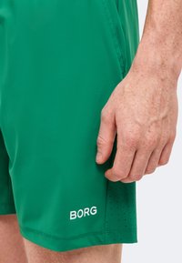Κοντινό πλάνο του χεριού ενός ατόμου που ακουμπά δίπλα σε πράσινα σορτς με διάτρητα πλαϊνά πάνελ και τη λέξη «BORG» τυπωμένη κοντά στο ρέλι.
