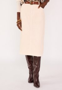 Jupe midi en velours côtelé beige clair avec poches latérales, coupe droite et ourlet brut. Associée à des bottes hautes bordeaux foncé.