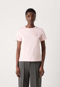 Rosa Baumwoll-T-Shirt mit kurzen Ärmeln und rundem Ausschnitt, mit einem strukturierten Logodetail auf der Brust, kombiniert mit grauen, gefältelten Hosen.