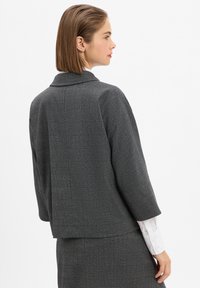 Grau strukturierte Jacke mit dezentem Karomuster, einreihigem Design und Kragen, kombiniert mit einer weißen Bluse darunter.