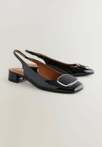  Černé lakované slingback boty se čtveratou špičkou, nízkým blokovým podpatkem a velkým kovovým akcentem v oblasti špičky.