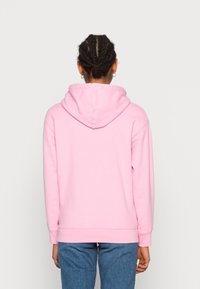 Persona con capelli intrecciati che indossa orecchini a cerchio, felpa rosa e pantaloni blu, vista da dietro su uno sfondo chiaro e semplice.