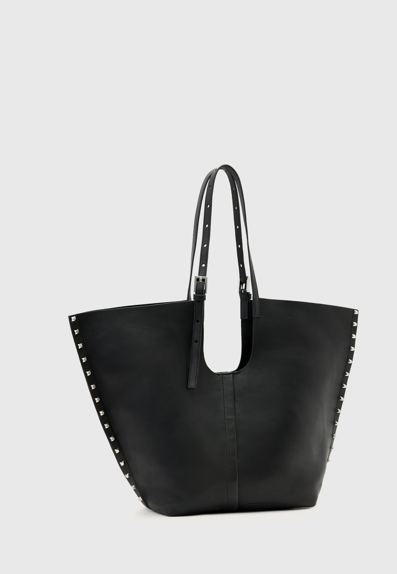 Borsa tote in pelle nera con borchie piramidali argento lungo le cuciture laterali e due sottili tracolle regolabili su sfondo grigio chiaro.