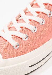 Koralowe płócienne trampki z białymi sznurówkami, srebrnymi oczkami i białą gumową podeszwą, z teksturowaną powierzchnią i wzmocnionym noskiem.