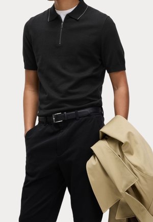 Mann, der ein schwarzes kurzärmeliges Poloshirt mit Reißverschluss, schwarzen Gürtel und schwarze Hose trägt, hält einen beigen Trenchcoat über dem Arm vor einfarbigem Hintergrund.