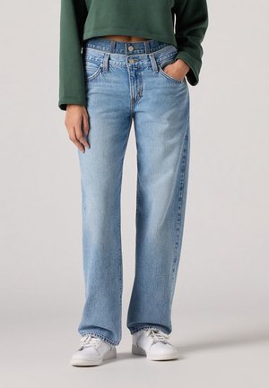 Kobieta ubrana w jasnoniebieskie jeansy z wysokim stanem i szerokimi nogawkami z podwójnym zapięciem na guziki, białe trampki oraz ciemnozielony krótki sweter.