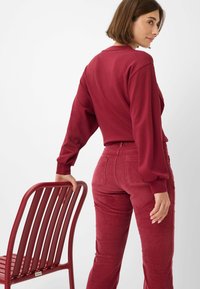 Bordeaux lange mouwen top met een relaxte pasvorm, gecombineerd met bijpassende corduroy broek. Een rode metalen stoel staat op de achtergrond.