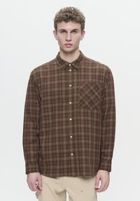 Chemise à carreaux marron en tissu, avec col boutonné, poche poitrine et accents beiges. Elle présente une texture douce et une coupe décontractée.