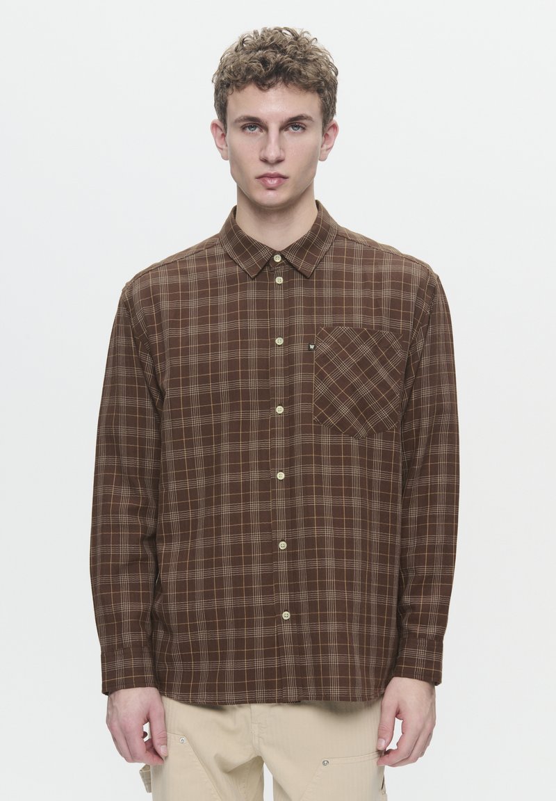 Chemise à carreaux marron en tissu, avec col boutonné, poche poitrine et accents beiges. Elle présente une texture douce et une coupe décontractée.
