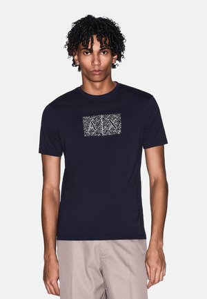 Giovane uomo con capelli ricci che indossa una T-shirt nera a maniche corte con un motivo geometrico bianco sul petto, abbinata a pantaloni beige chiaro.