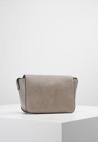 Obdélníková taupe kožená kabelka přes rameno s klopovým uzávěrem, černým lemem a minimalistikým designem, vystavená na bílé podstavě.