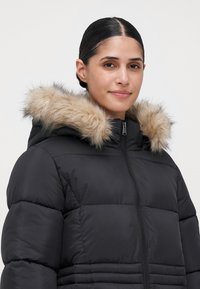 Mujer con cabello oscuro, vestida con una chaqueta de plumas negra con capucha de pelo, de pie frente a un fondo claro y liso, mirando hacia el frente.