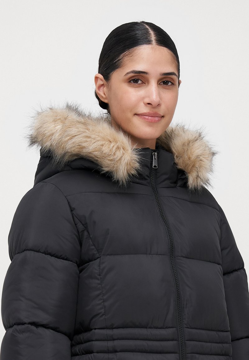 Mujer con cabello oscuro, vestida con una chaqueta de plumas negra con capucha de pelo, de pie frente a un fondo claro y liso, mirando hacia el frente.