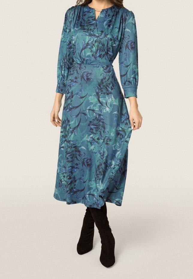 Lang teal kjole med blomstermønster, der har V-udskæring, lange ærmer og en løs silhuet. Sammen med sorte knæhøje støvler.