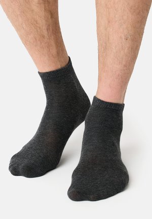 Ben med lyst hår, iført mørkegrå ankelsokker, stående på en ensfarvet hvid baggrund.