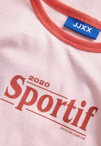 Světle růžové tričko s červeným kulatým výstřihem. Na přední straně je nápis "Sportif 2020" tučnými červenými písmeny, pod ním se nachází "ATHLETIC AND LEISURE CLUB".
