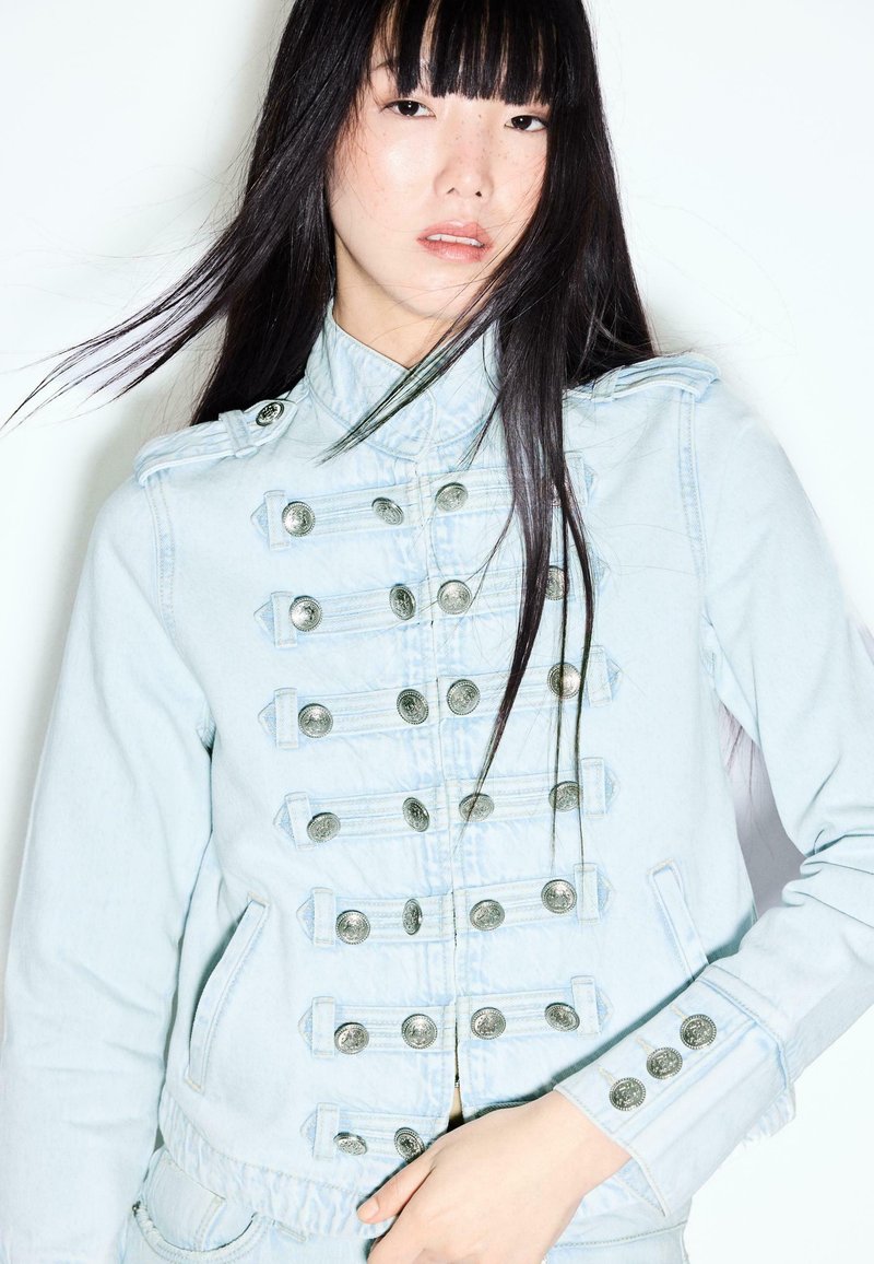 Femme aux longs cheveux noirs porte une veste en jean bleu clair style militaire avec plusieurs boutons argentés, debout sur fond blanc.
