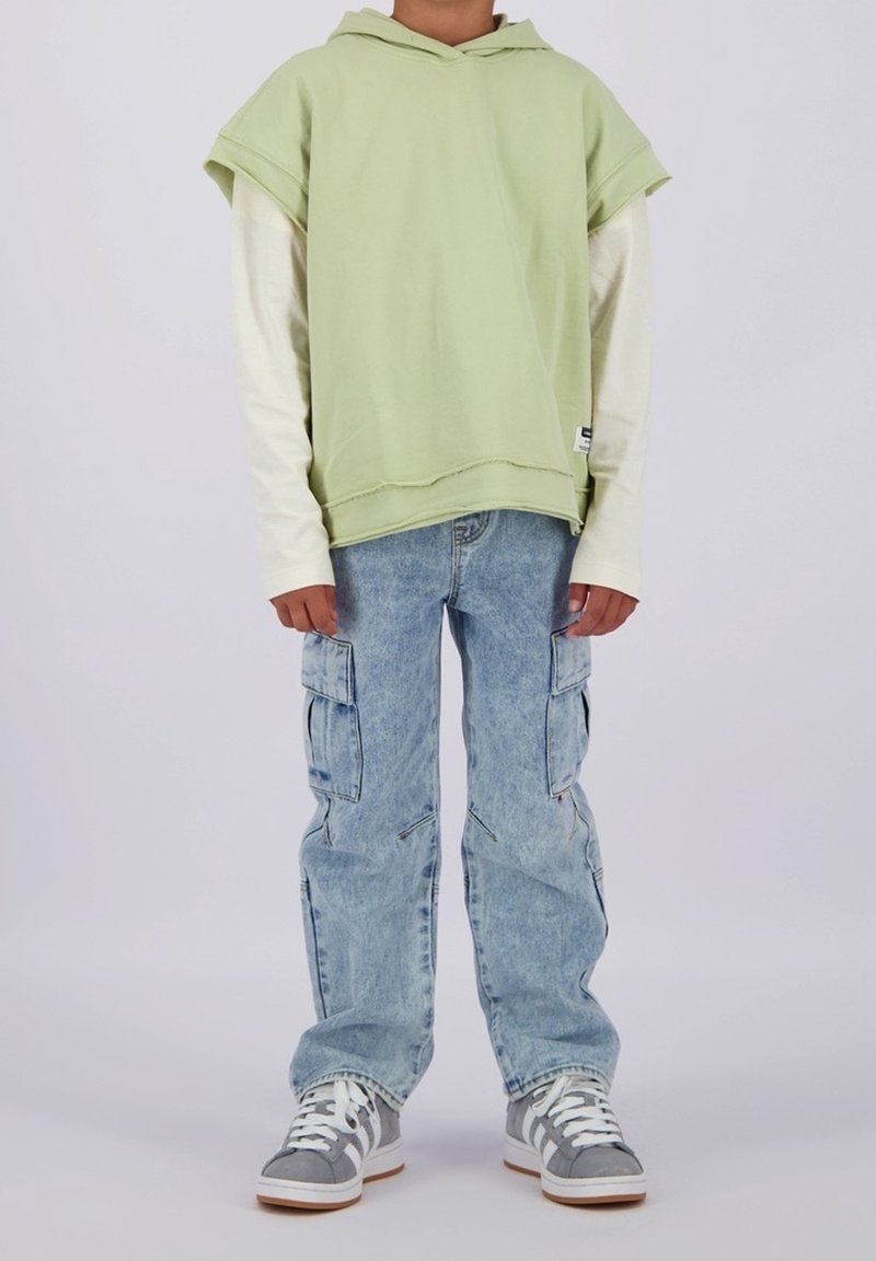 Enfant portant un sweat à capuche sans manches vert clair sur un t-shirt blanc à manches longues, un jean cargo bleu clair et des baskets grises, debout devant un fond uni.