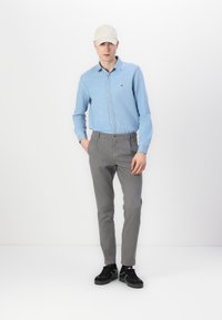 Chemise en denim bleu clair, pantalon slim gris, baskets noires et casquette beige. La tenue présente un logo discret sur la chemise.