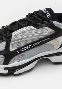 Czarno-srebrny but sportowy z siateczkową cholewką, wyściełanym językiem oraz wyraźnym brandingiem "Lacoste" po boku i pięcie. Teksturowane detale na całej powierzchni.