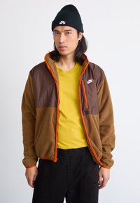 Chaqueta de forro marrón con acentos naranjas, bolsillos con cremallera y parte superior de nailon marrón. Usada sobre una camisa amarilla y combinada con pantalones negros.