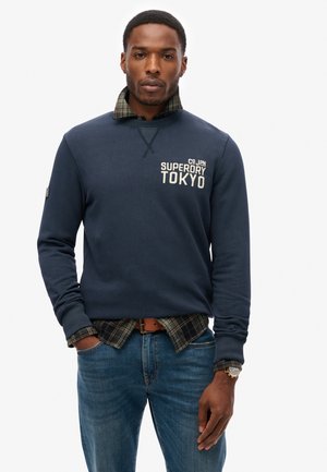 Marineblaues Sweatshirt mit dem Aufdruck "Superdry Tokyo", über einem karierten Hemd getragen. Passt gut zu Jeans und einem braunen Gürtel. Uhr am Handgelenk.