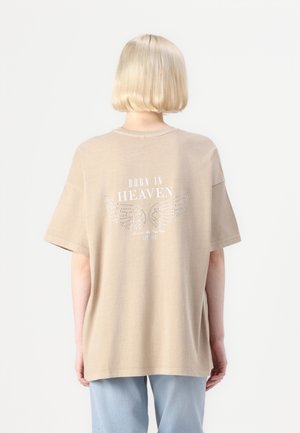T-shirt beige surdimensionné avec un design imprimé de ailes et le texte "NÉ AU PARADIS", sur fond uni.