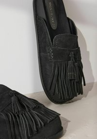 Mules en suède noir avec un accent de franges et un pompon décoratif. Le design comprend un bout arrondi et une semelle plate. La texture est douce.