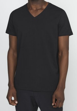 Mand iført en enkel, sort T-shirt med V-hals og korte ærmer samt mørke bukser, vist fra nakken til hofterne mod en lys baggrund.