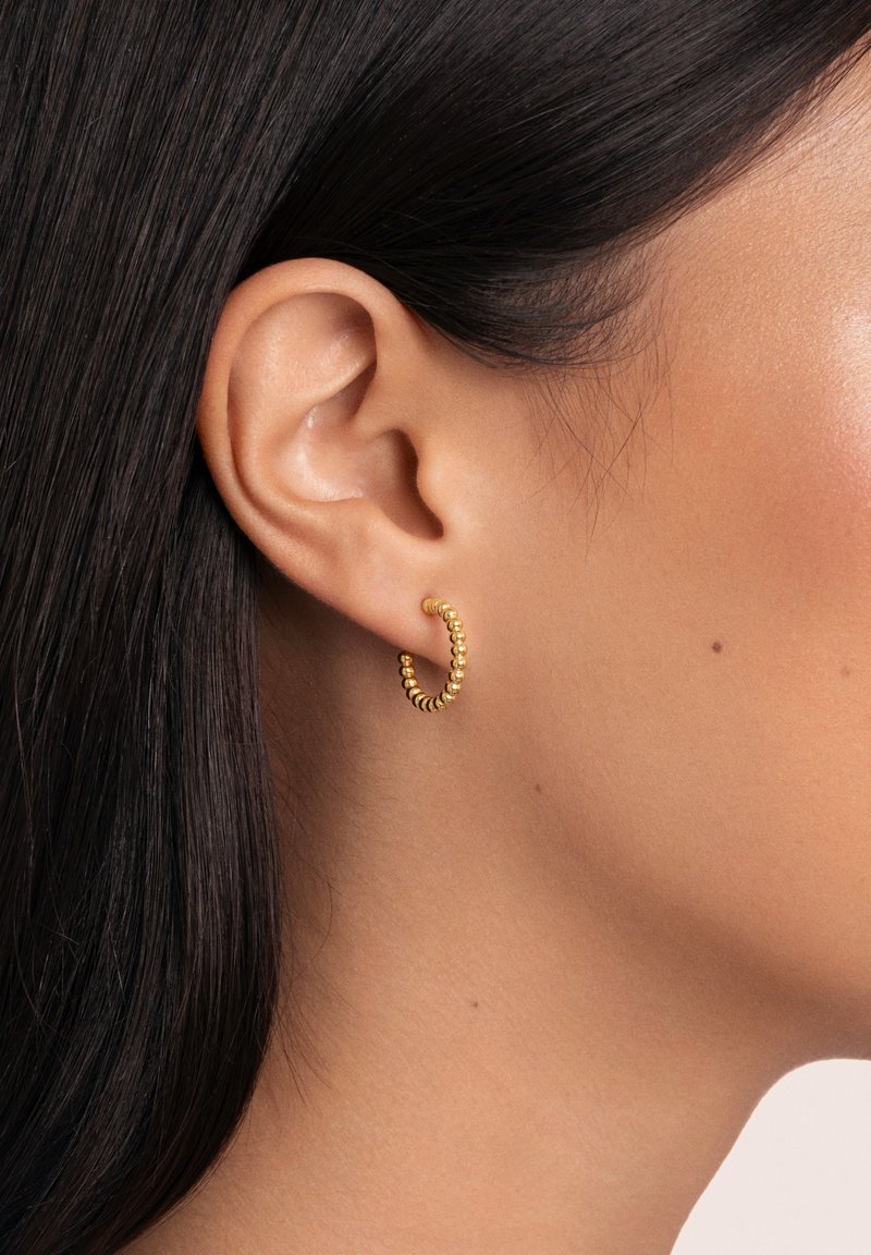 Pendiente de aro en oro con textura de cuentas, forma circular y acabado pulido. Llevado en una oreja perforada junto a un cabello liso y oscuro.