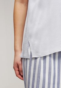 Světle šedá košile s jemnou strukturou, s bočními rozparky. Kombinováno s oděvem, který má horizontální modro-bílé pruhy.