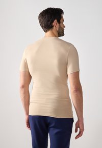 Cămașă bej cu mânecă scurtă, cu un design ajustat, textură netedă și decolteu rotund, purtată cu pantaloni navy, arătând din spate.