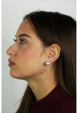 Joven mujer de cabello largo y oscuro que lleva pendientes de perlas, mirando de lado contra un fondo claro y liso, vestida con una camiseta granate.